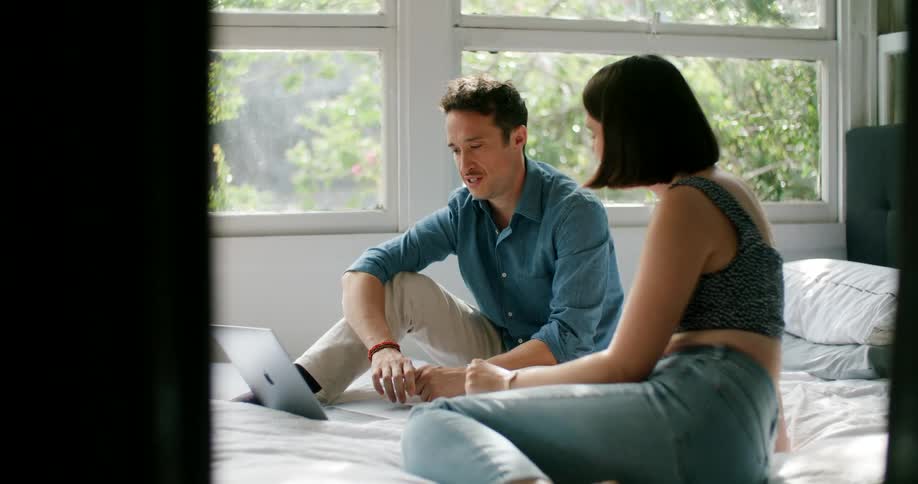 A man and woman are sitting on a bed together with a laptop