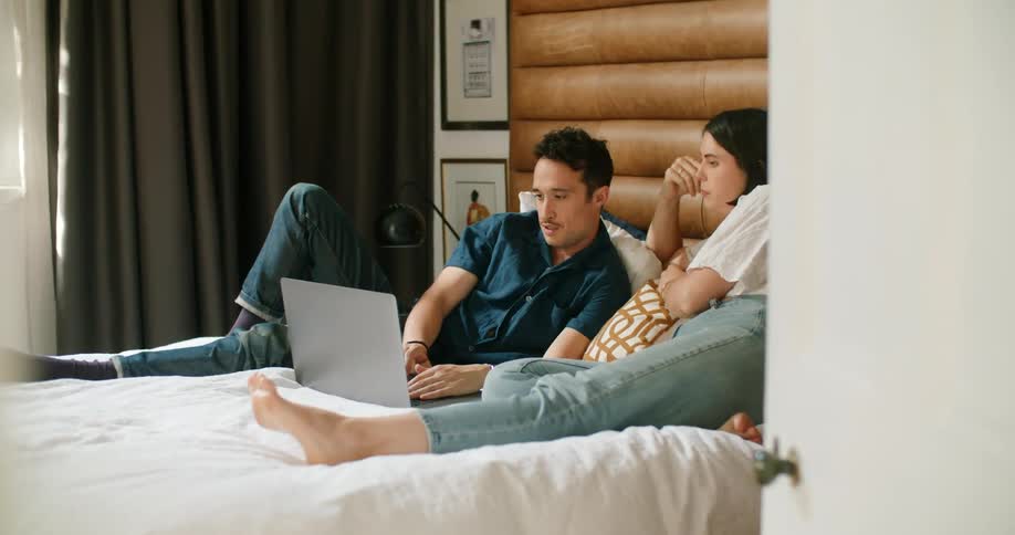A man and woman resting together on a bed speak to one another as they gaze at a laptop lying on the bed