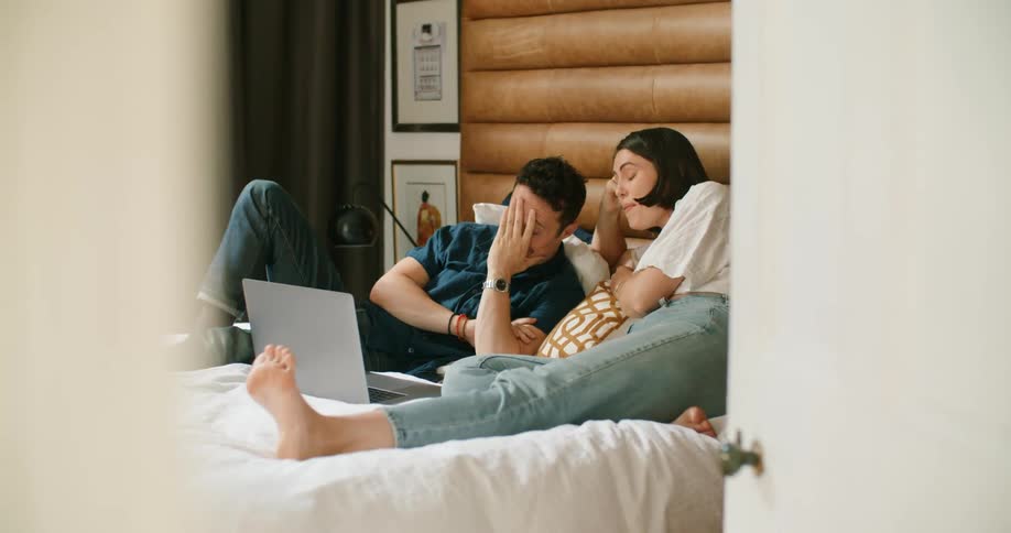 A man and woman are laying on a bed and looking at a laptop