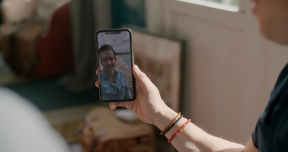 Two people sit at home and video chat with a man on a beach on a cell phone