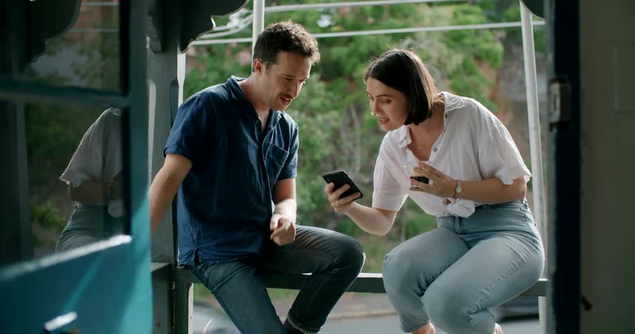 A woman sitting on the railing of a balcony shows her smartphone to a man sitting next to her