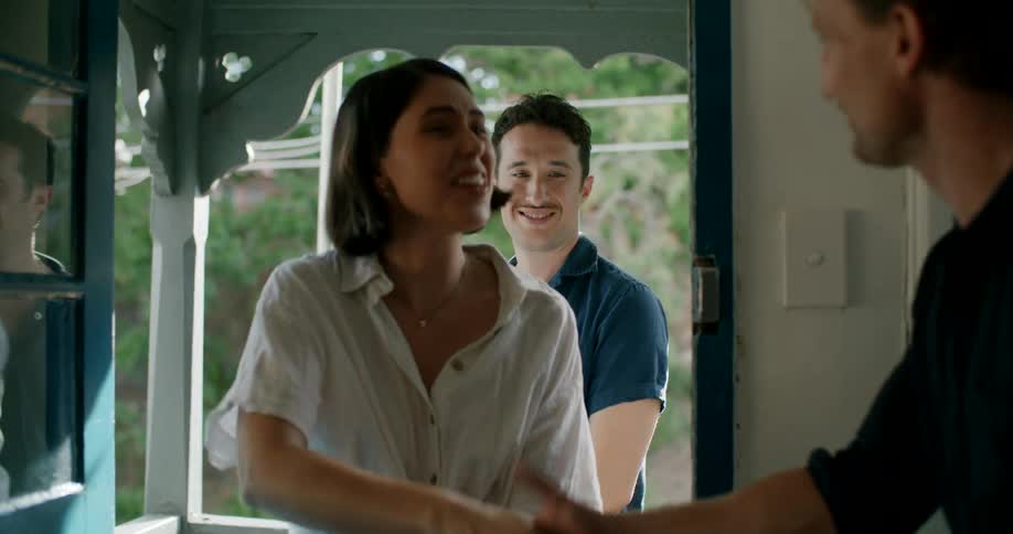 A man opening a door and inviting a man and a woman to enter into a house and shaking their hands