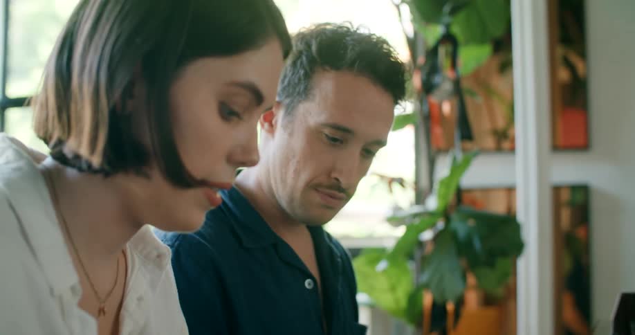 A man and a woman in a room sitting at a piano with a window in the background