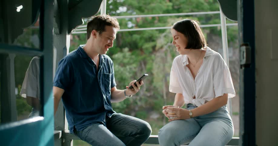 A couple sit on a railing drinking coffee and laughing at what they share on a phone