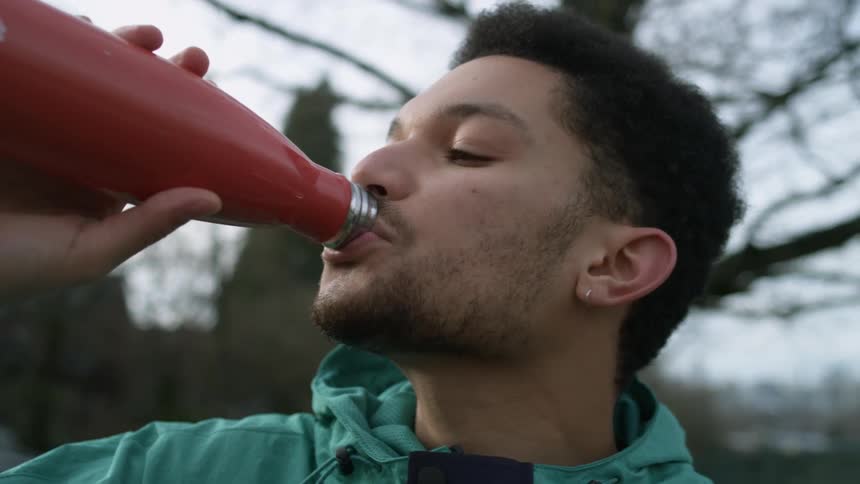 A black male teenager stands outdoors in a neighborhood taking a drink from a red bottle