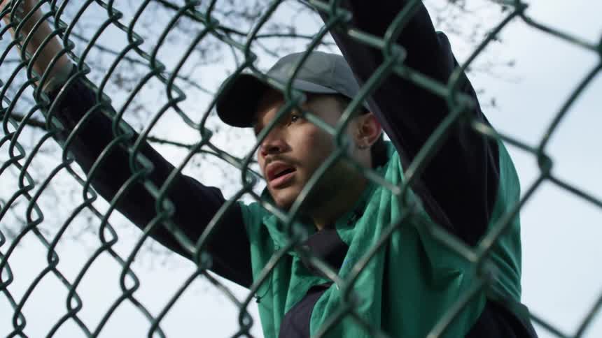 Young man wearing sports clothes stops for a moment to recline on a wired fence before continuing excercising