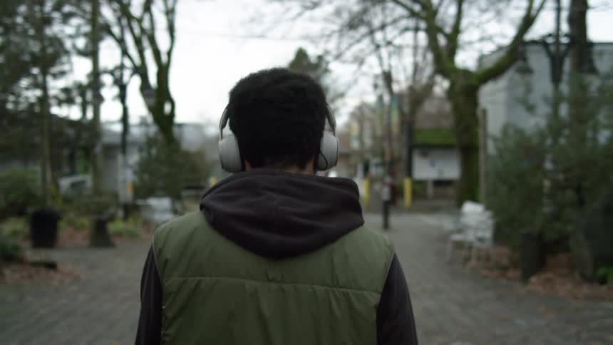 A man lifts his headphones from around his neck to his ears as he walks along a walkway through a town during the day