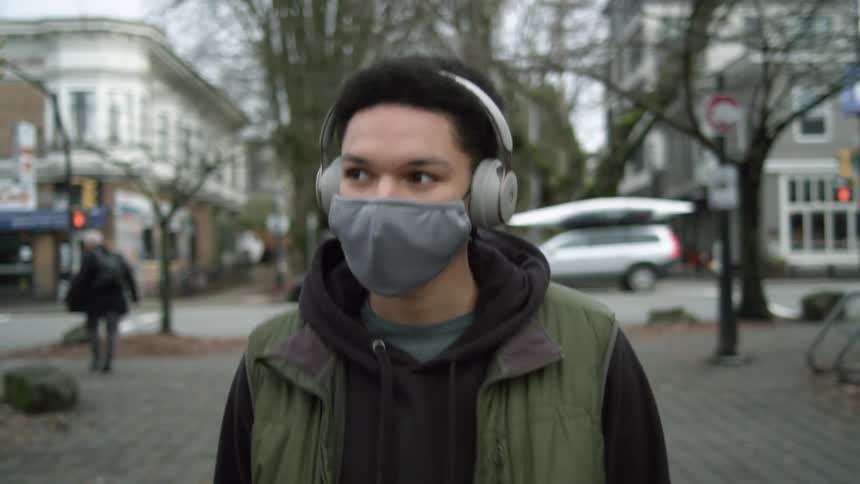 A male teenager walks on a sidewalk in a small town wearing headphones and a face mask