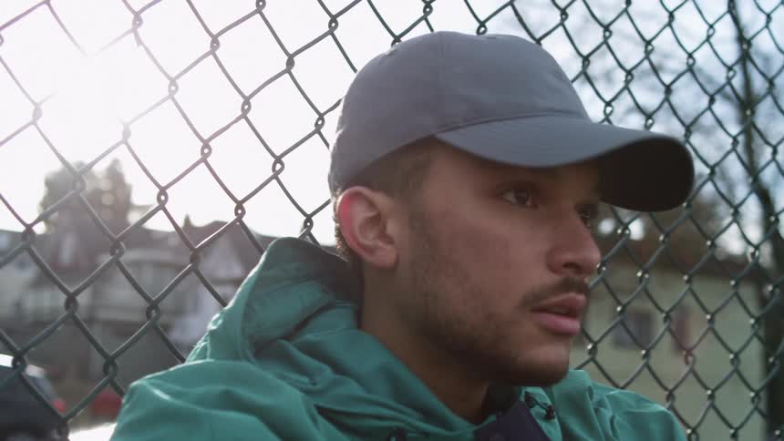 A man with a hat and a jacket sitting by a fence with vehicles buildings the sky and clouds in the background