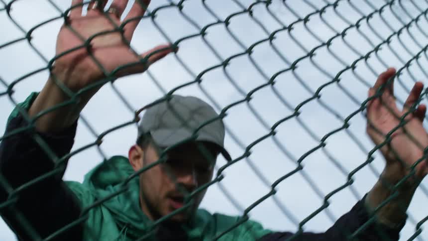 A man with a jacket and a hat running up to a fence and running back with buildings the sky and clouds in the background