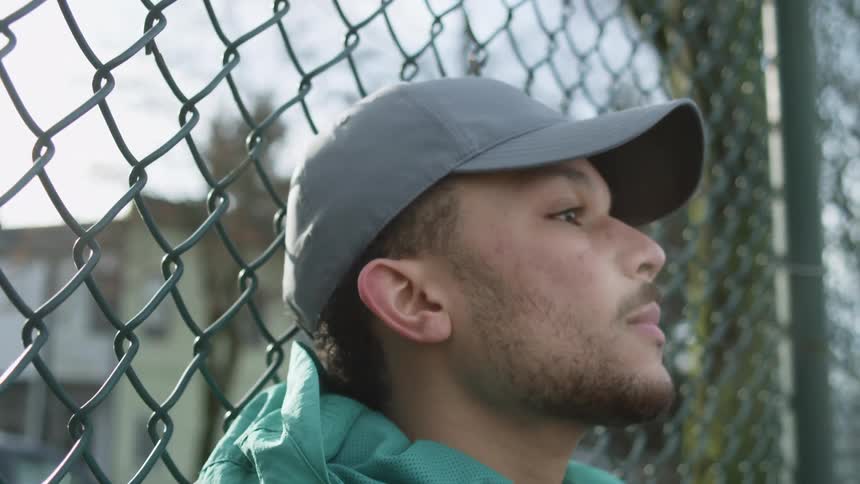 Tired young man sitting on the ground reclined on a wired fence in a soccer field at morning