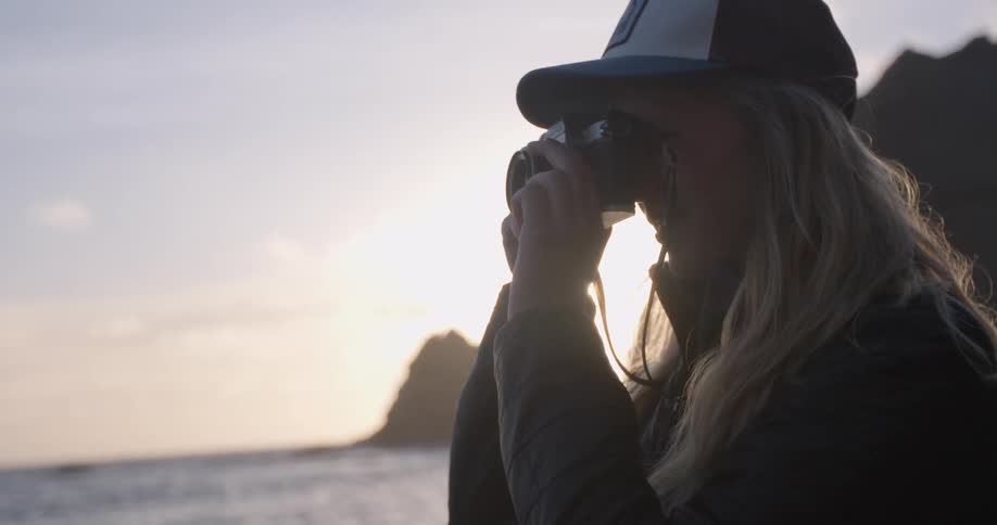 Blonde woman in a baseball cap taking a picture of the ocean at sunset