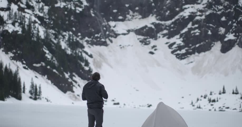 A person standing near a tent takes a picture of a snow covered mountain on a cold winter day