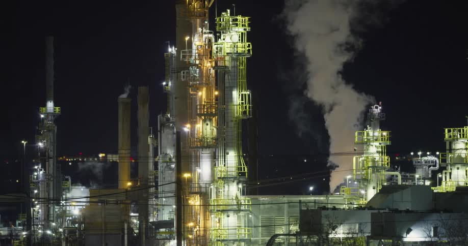 A factory with lights chimneys and smoke with the sky in the background