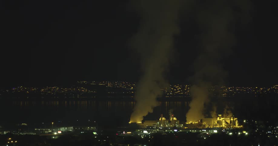 An aerial view of smoke billowing out of factory on the coast and a city skyline in the distance