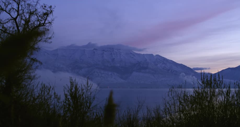 Clouds over the snow covered peaks of a mountain range near a lake at sunrise