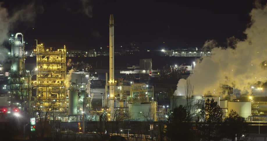 A factory with lights chimneys and smoke with the sky in the background