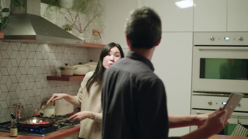 A woman cooking in a skillet in a home kitchen speaks to a man holding a tablet next to her