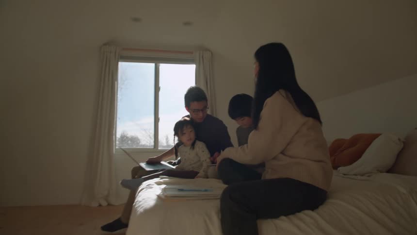 A family sits together on a bed looking as a boy uses a tablet