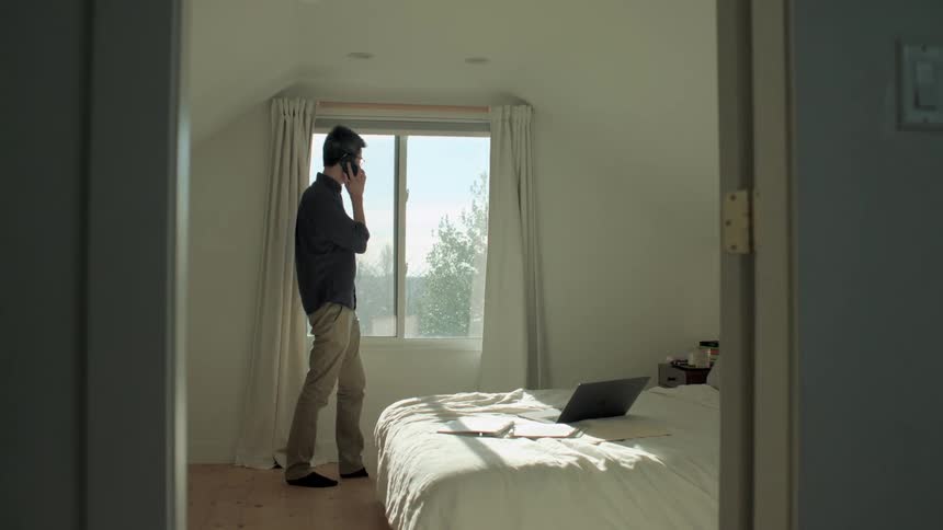 A man answering a cell phone while pacing in front of a bedroom window