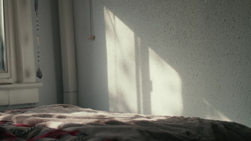 An empty room with a blanket on a bed and an open window with sunlight streaming through