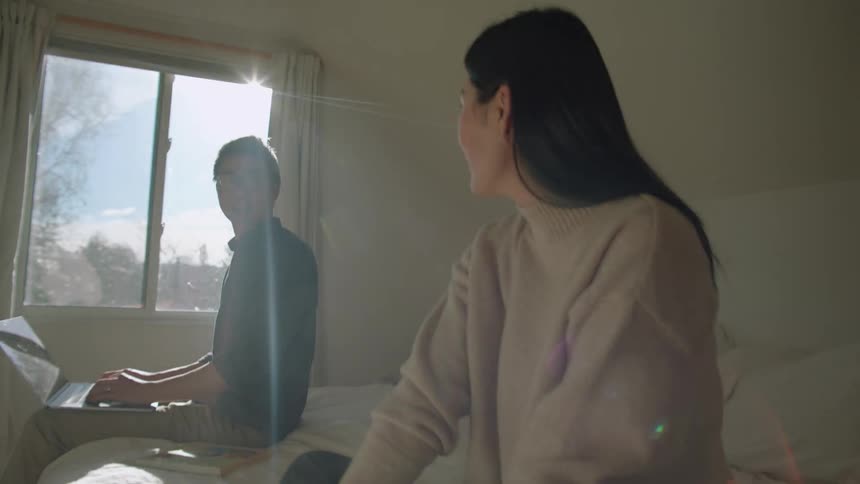 A woman in a bedroom sitting on a bed talking to a man typing on a laptop