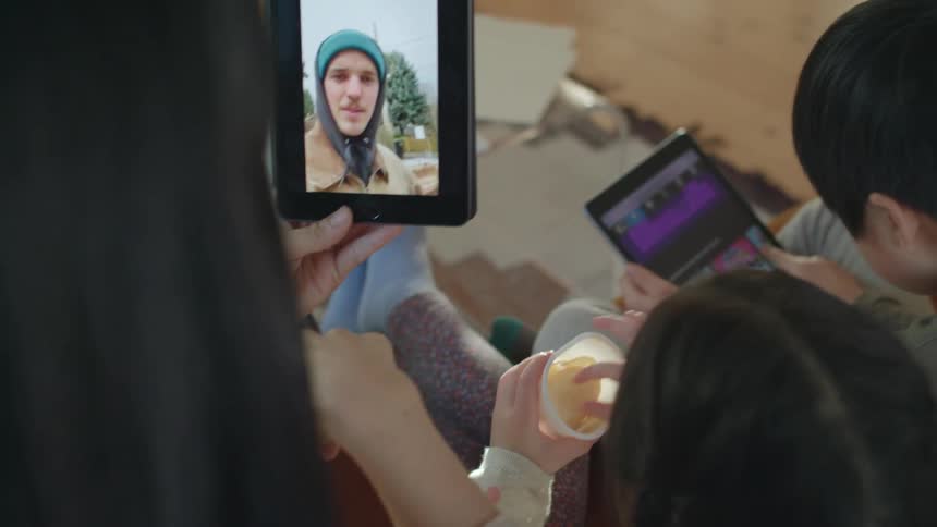 A woman talks to her son on a tablet while sitting inside with two other children
