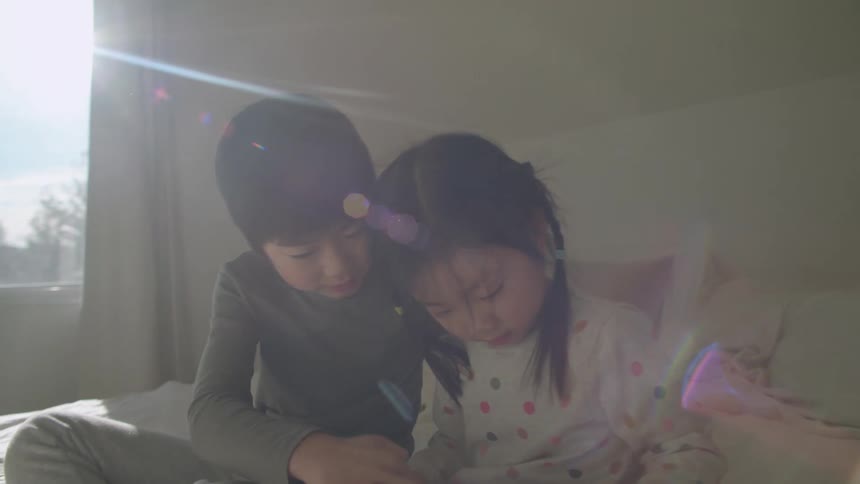 A young boy and girl play with a computer tablet while sitting together on a bed on a sunny day