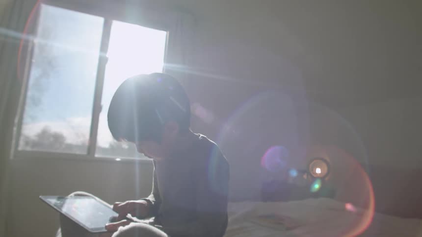 A boy sitting on a bed using a tablet with a wall and window in the background