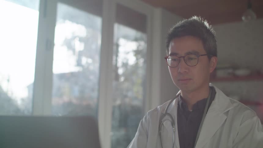 A physician is sitting indoors at his computer while typing
