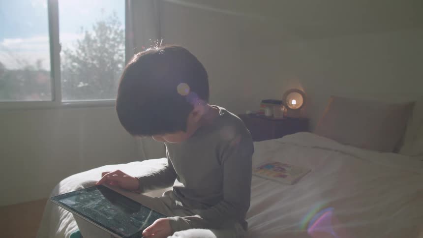 A little boy plays on a tablet while sitting on a bed during the day