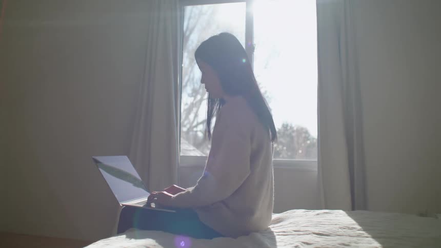 Young woman working on a laptop sitting in the bed at home answers a call on her smartpone