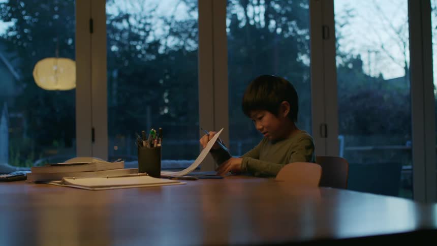 A boy in a room sitting at a table holding a pen and looking through papers