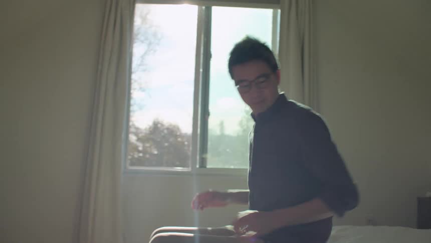 A man at home looks at his cell phone and then sets it on the bed which he sits on and picks up his laptop