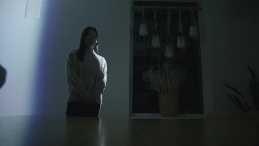 A woman standing in a room next to a table and a chair with a window and plants in the background