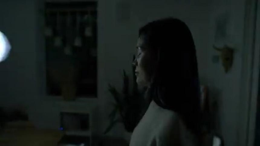 A woman stands in her living room with her eyes closed as a passing vehicle shines its headlights through a window