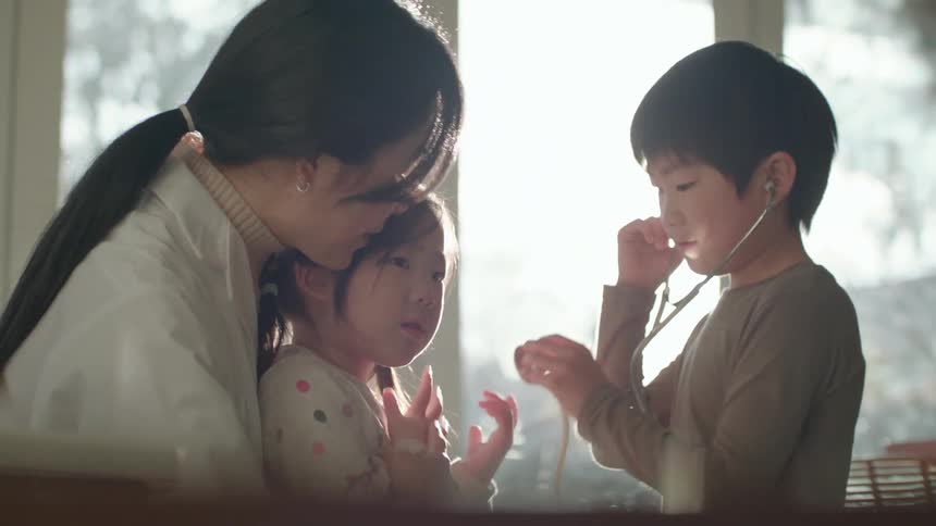 A woman sitting on a chair with a girl sitting on her lap as a boy listens to the girls heart with a stethoscope