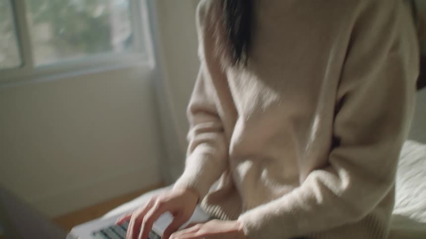 An asian woman sits on a bed typing on a laptop on her lap