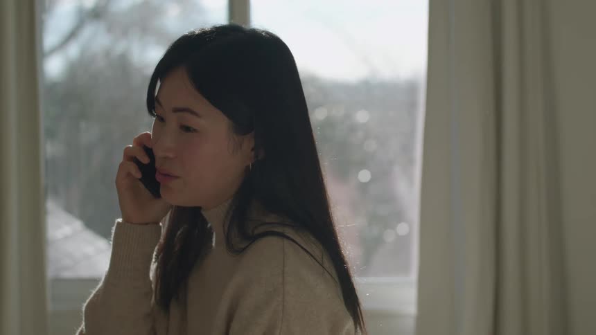 A woman talking on a cell phone in front of a sunny window at home