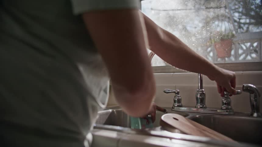 A man stands at a kitchen sink while washing a bowl that he places on the adjacent counter top