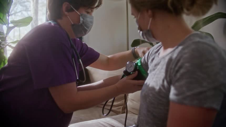 A woman with scrubs a mask and a stethoscope checking the blood pressure of a woman sitting on a couch