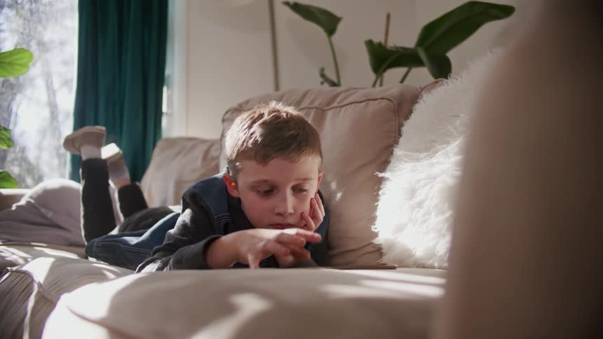 A young boy relaxes on a couch while playing on a tablet computer