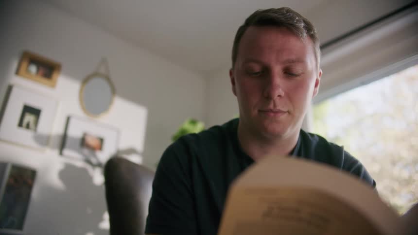 A man sits in a chair in his living room and leans forward while reading a paperback book