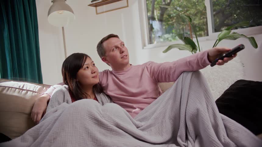 A man sits on a couch at home with his arm around a woman and presses buttons on a remote control as they find something to watch