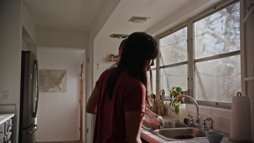 A woman hugs her husband from behind as he washes dishes in their kitchen and stops to help him dry them off