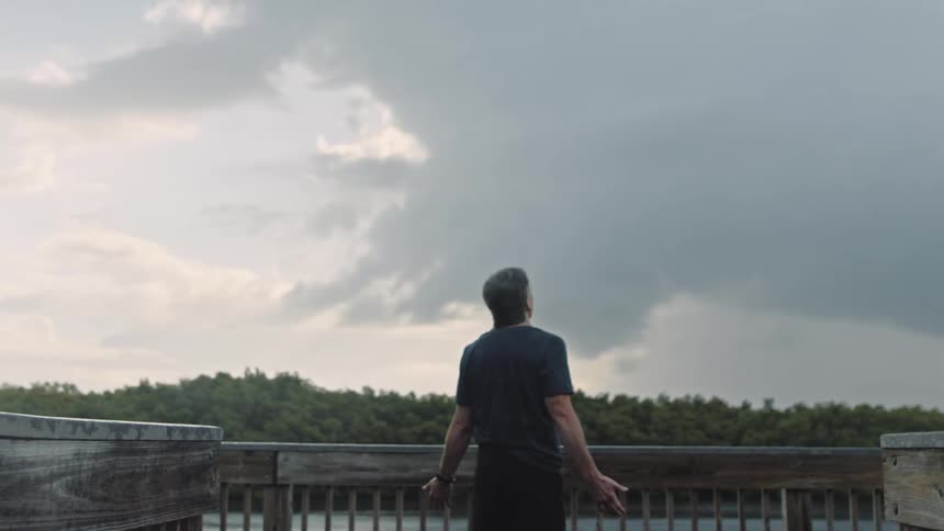 A man standing near a coast with trees the sky and clouds in the background