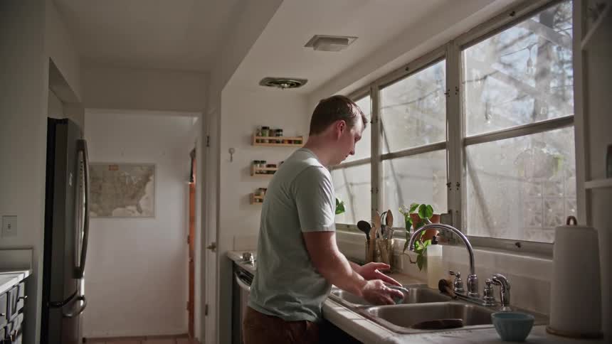 A man is doing dishes indoors next to a window
