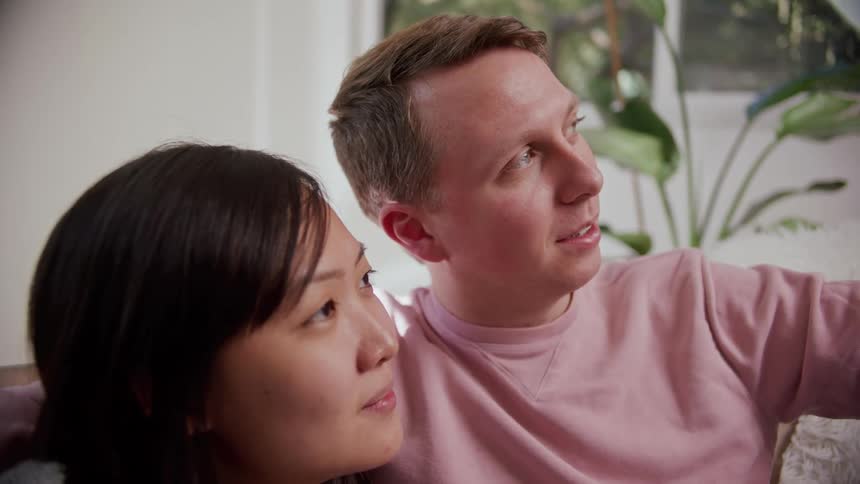A man sits with his arm around his girlfriend as they talk and watch television