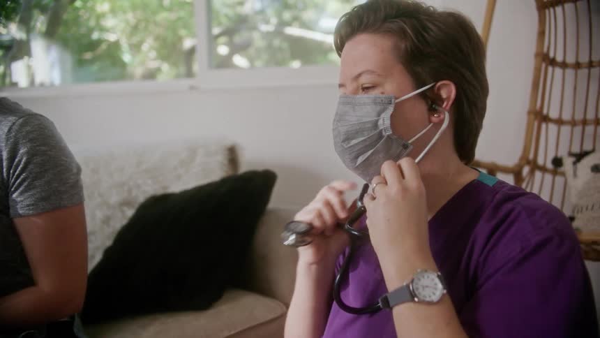 A nurse speaks with a patient at home and puts on a stethoscope to check her heartbeat