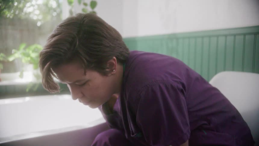 A woman sitting on a chair with scrubs putting on shoes in a bathroom next to a bathtub